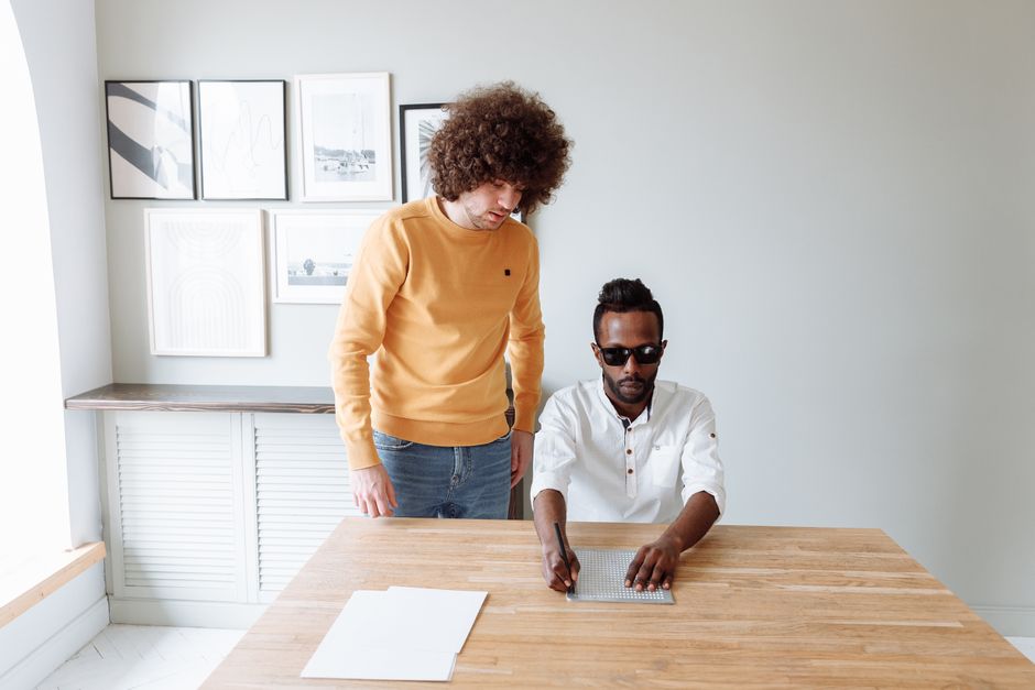 Mentor guides blind man using slate and stylus for Braille writing on a wooden table.